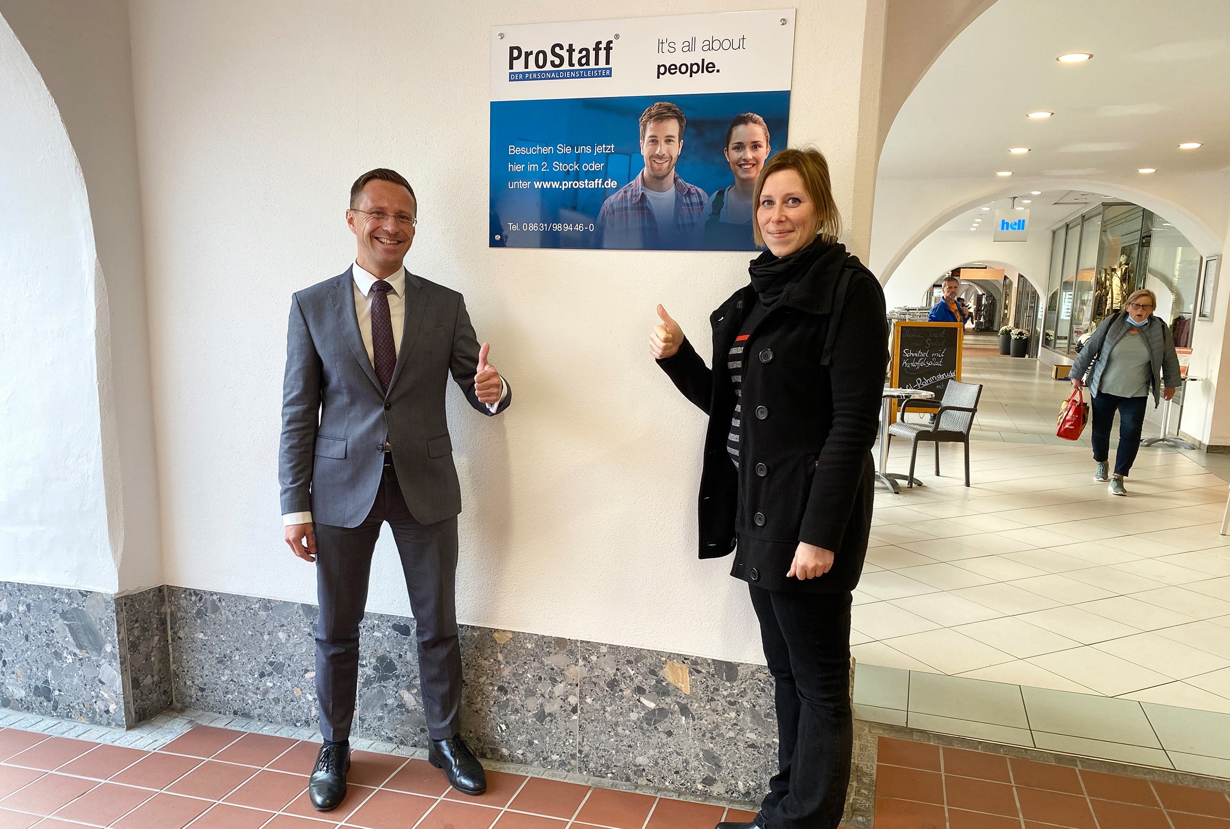 Zwei Mitarbeiter von ProStaff stehen lächelnd mit Daumen nach oben vor dem Schild des neuen Standortes in Mühldorf