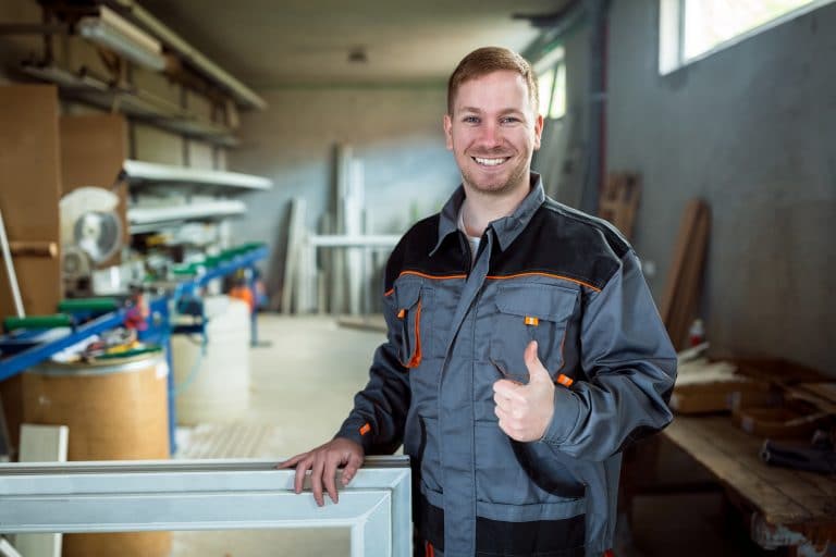 Mann in Arbeitskleidung steht in einer Werkstatt und lächelt mit Daumen nach oben in die Kamera